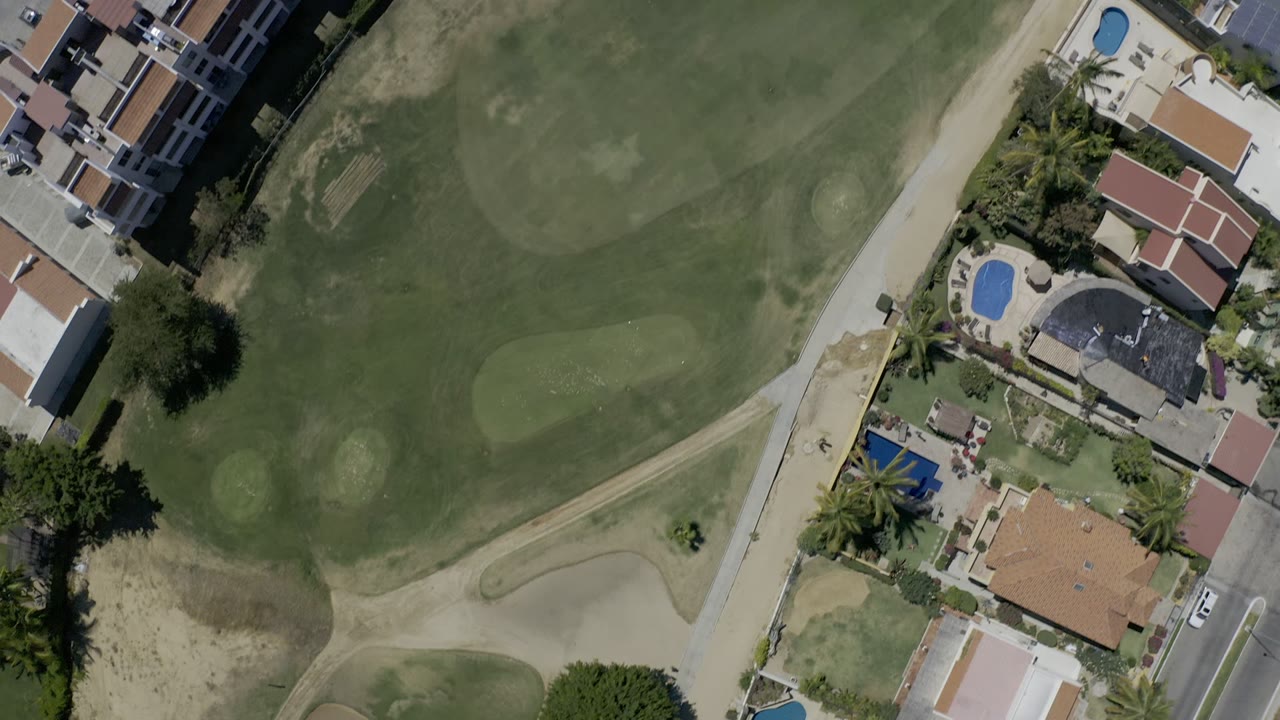 High overhead Los Cabos aerial with fairway greens across the frame and a residential strip with pools and tiled roofs along the right side, with no visible people.