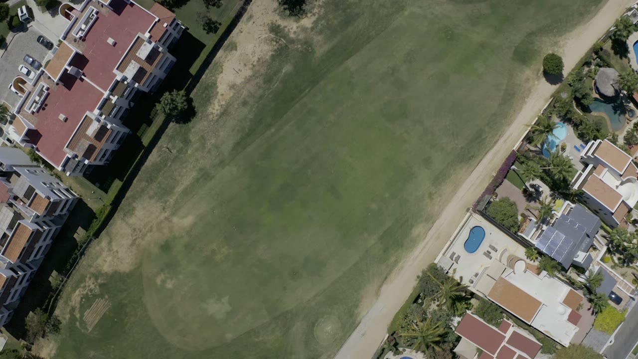 Opening overhead frame in Los Cabos, Mexico, showing a green golf fairway, tan path, red-roofed residences, small pools, palms, and no visible people.