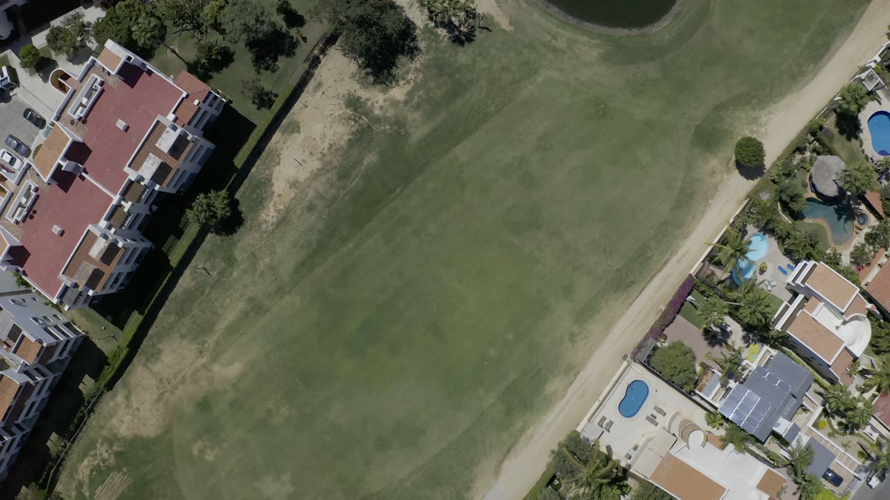 Top-down aerial frame in Los Cabos, Mexico, showing a green golf fairway bordered by large homes, pools, palm landscaping, and a pale road, with no visible people.