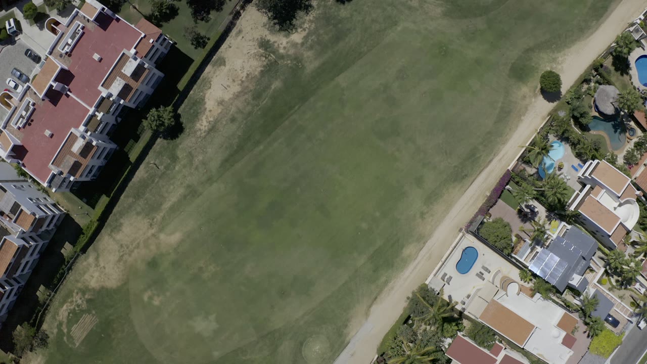 Aerial frame over Los Cabos, Mexico, with the fairway filling most of the view and residential buildings, blue pools, parked vehicles, and landscaping along one side, with no visible people.