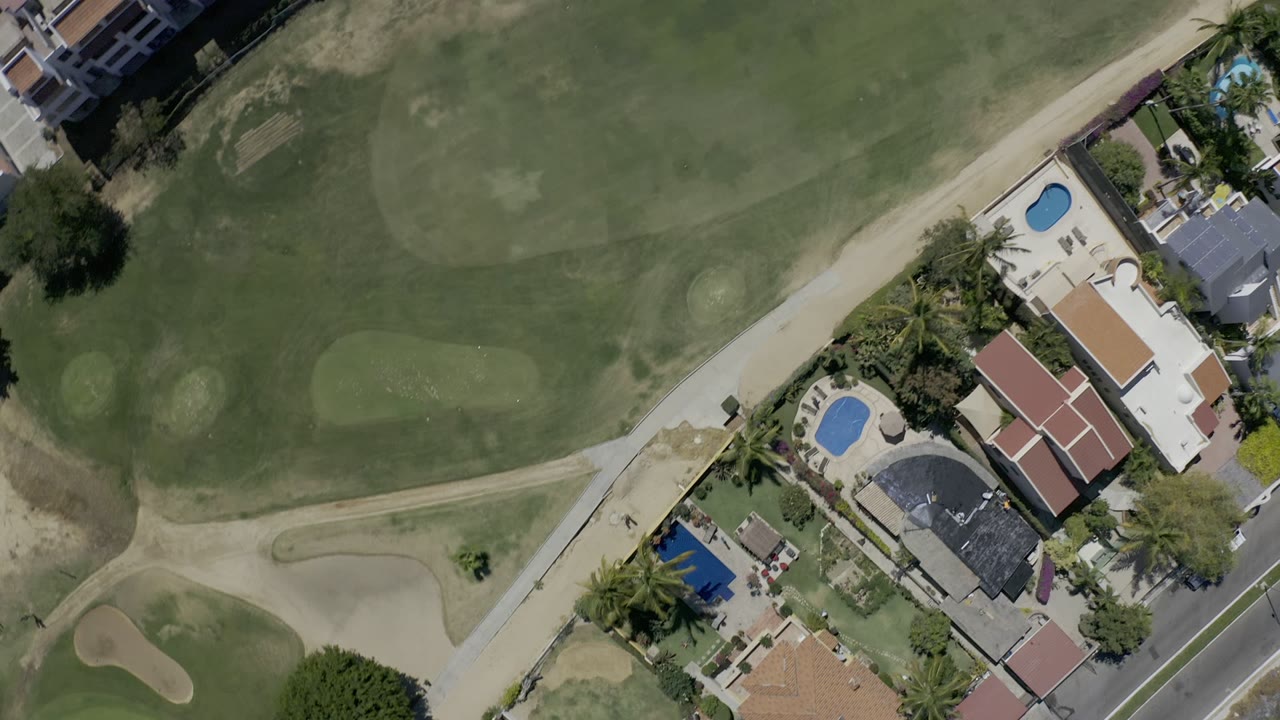Top-down aerial view in Los Cabos showing golf fairway grass, sand traps, cart paths, private homes, blue backyard pools, palms, and no visible people.