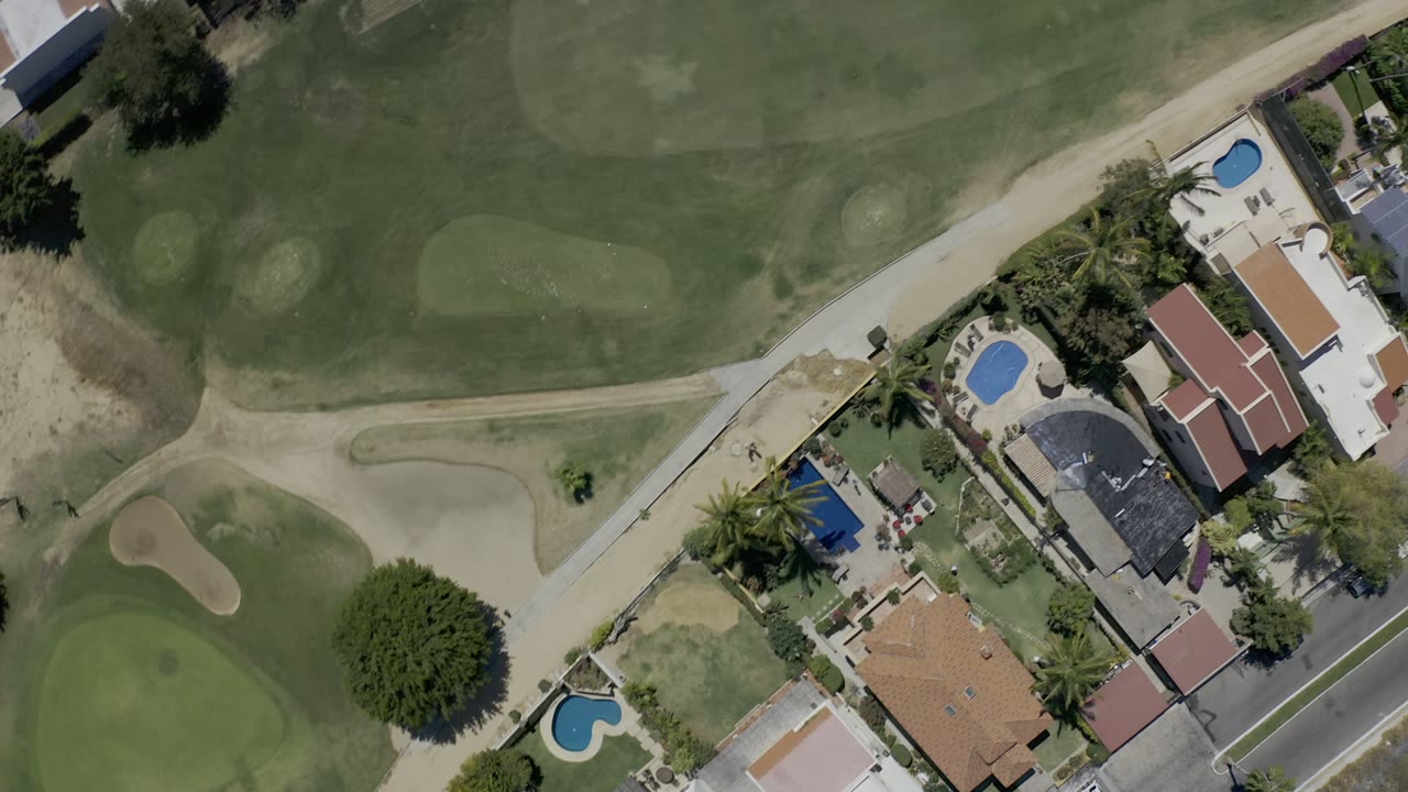 Overhead Los Cabos frame with manicured golf greens, pale sand bunkers, a course path, adjacent houses, pool patios, trees, and no visible people.