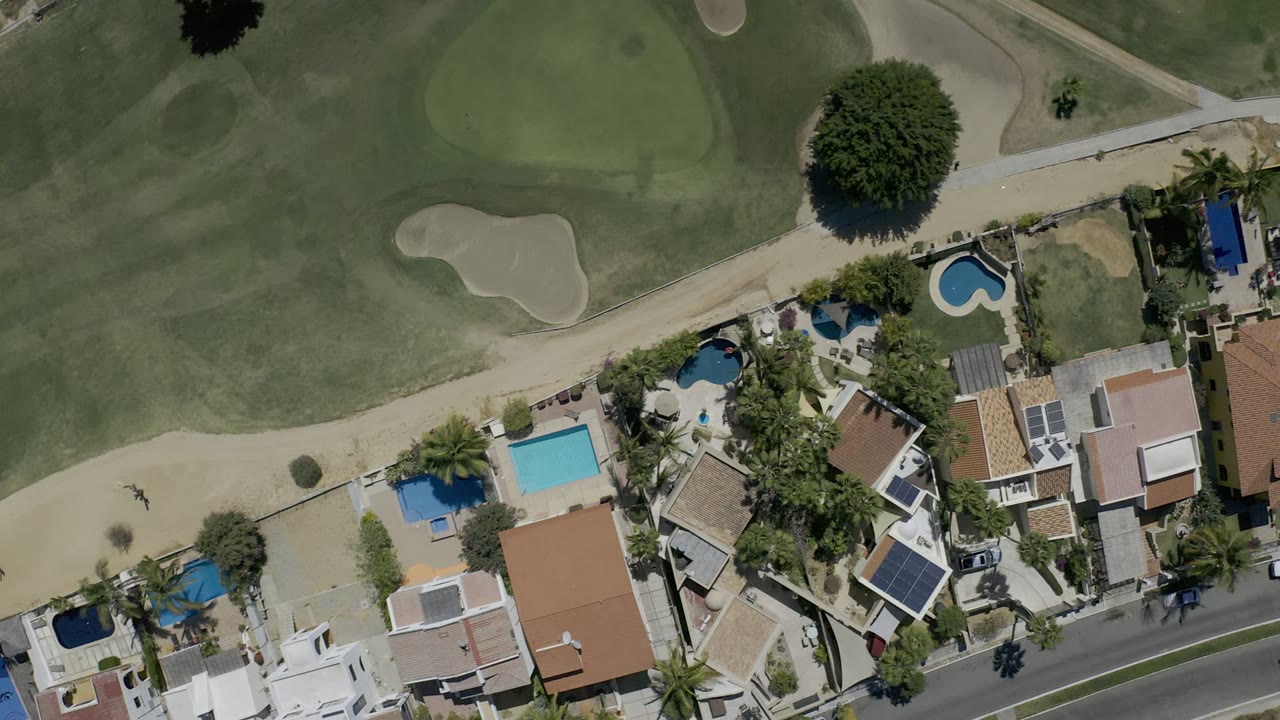 Overhead Los Cabos frame with the golf course across the top, a pale path through the middle, and clustered homes with pools below, with no visible people.