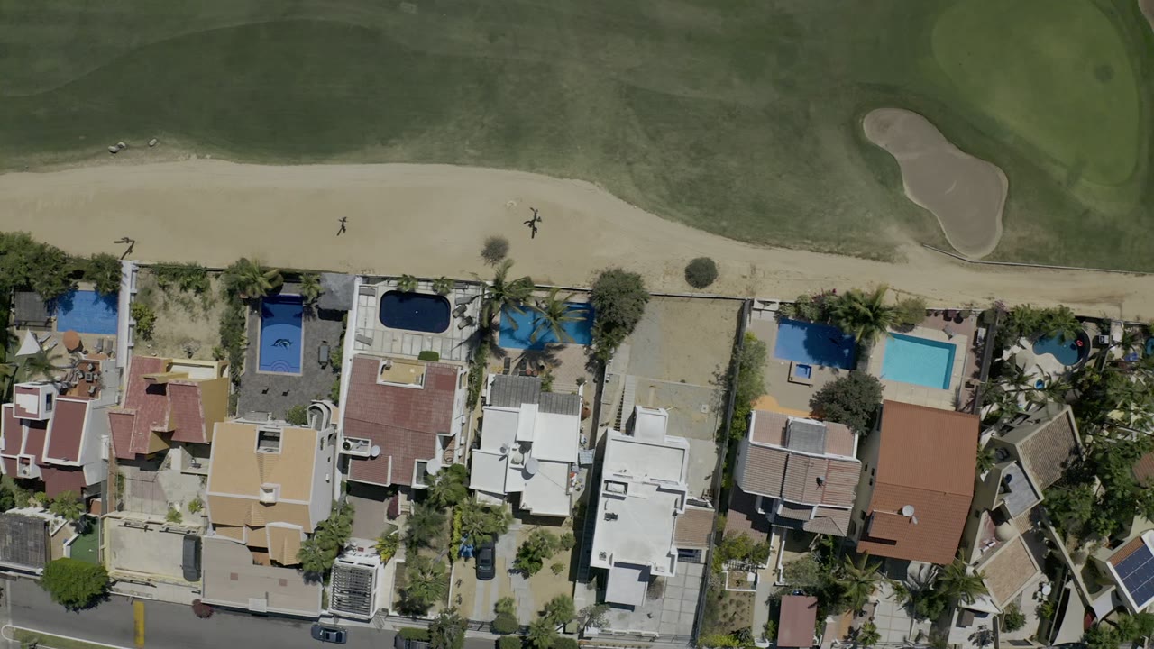 Overhead Los Cabos frame showing private homes, blue pools, palm trees, a road, and a golf-course sand bunker at the top, with no visible people.