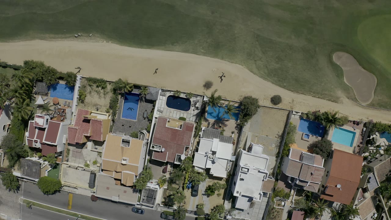 Top-down Los Cabos aerial with red and white roofed houses, several private pools, a curved road, and fairway grass with a sand trap, with no visible people.