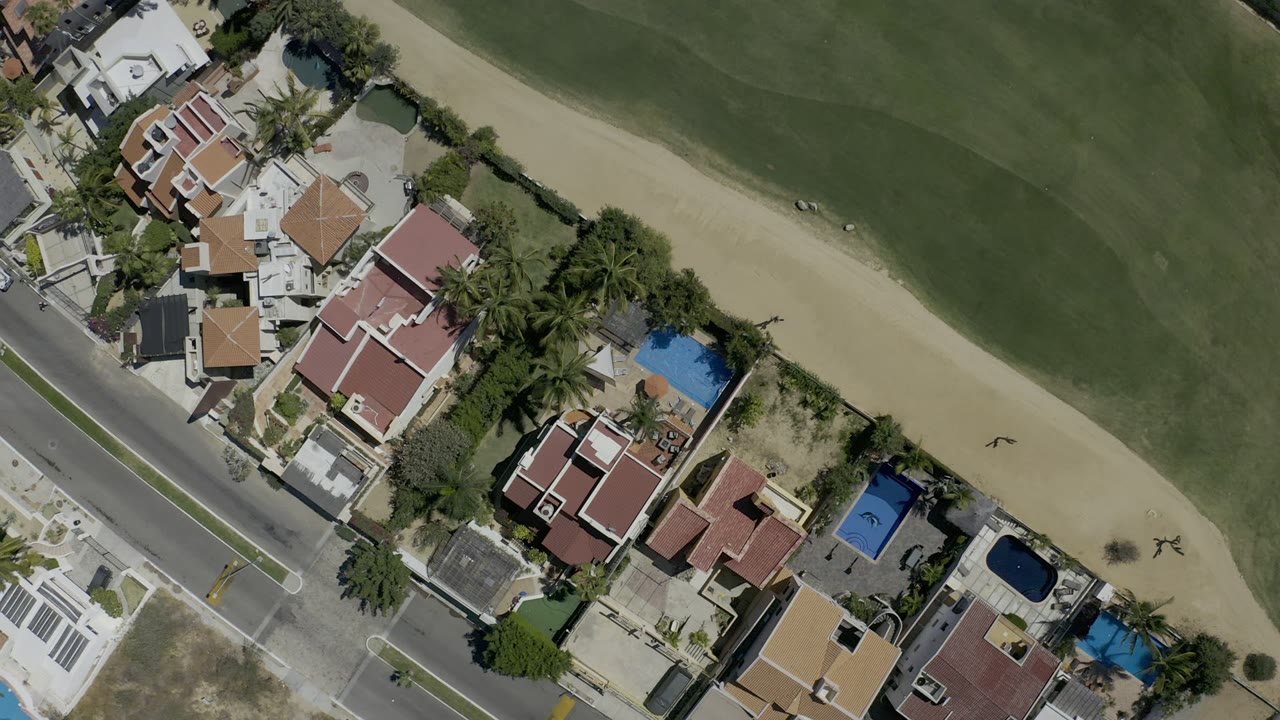 Overhead Los Cabos view with clustered residential roofs, walled yards, palm trees, small blue pools, a paved road, tan sand, and broad green course area with no visible people.