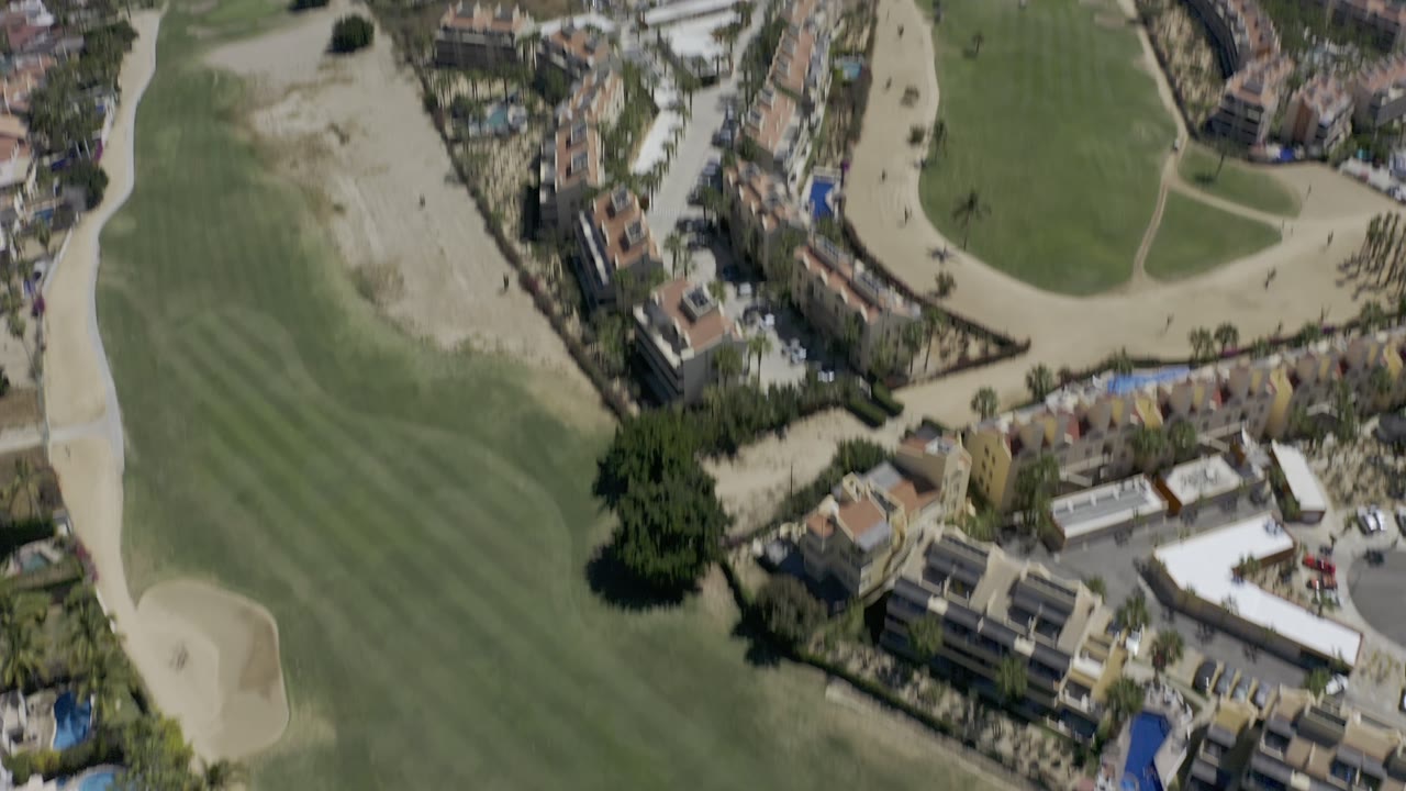 Aerial frame over Los Cabos, Mexico, with golf greens and sand areas in front, clustered buildings near the center, coastline beyond, and no visible people.