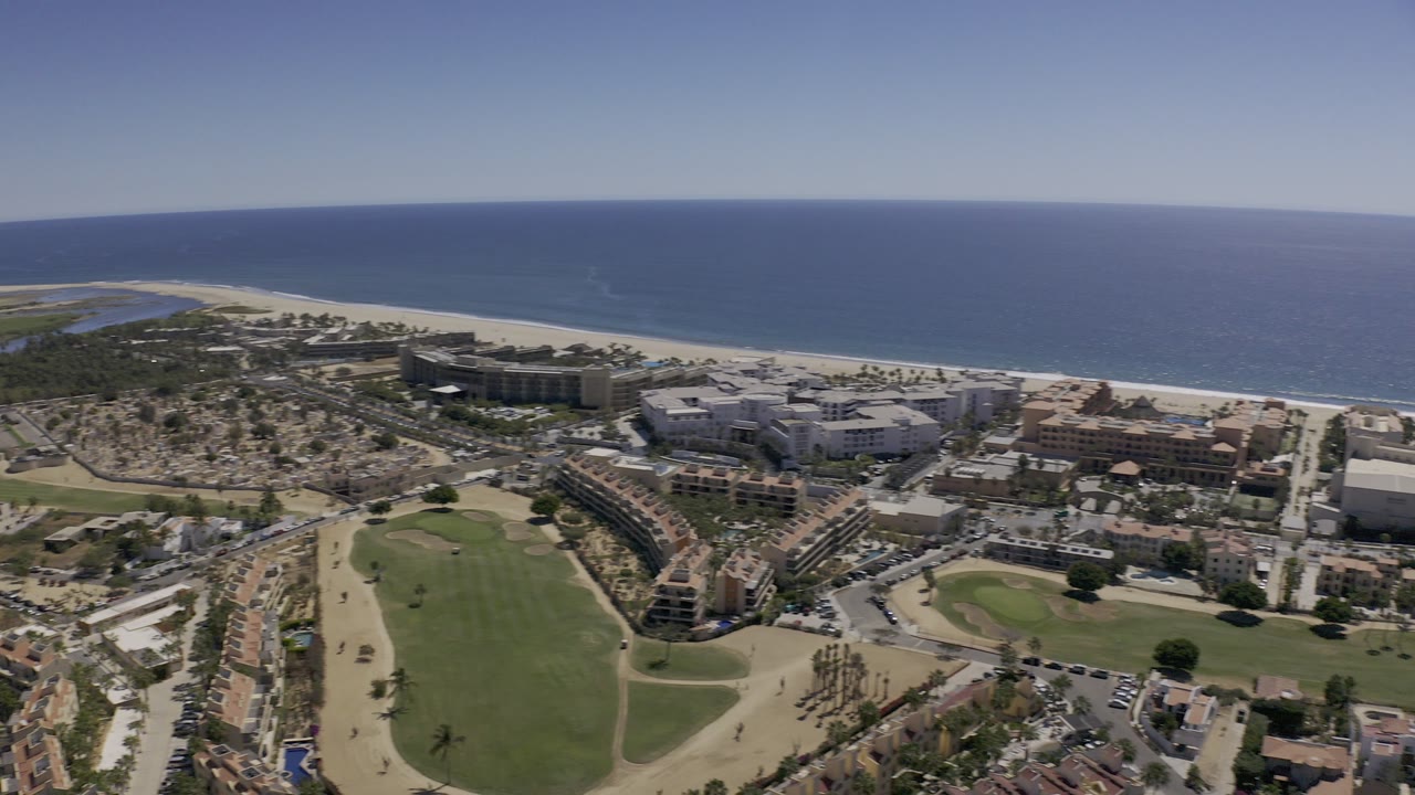 Aerial view over Los Cabos, Mexico, with blue ocean beyond a pale beach, beachfront buildings, roads, palms, and a large green open area in the foreground, with no visible people.