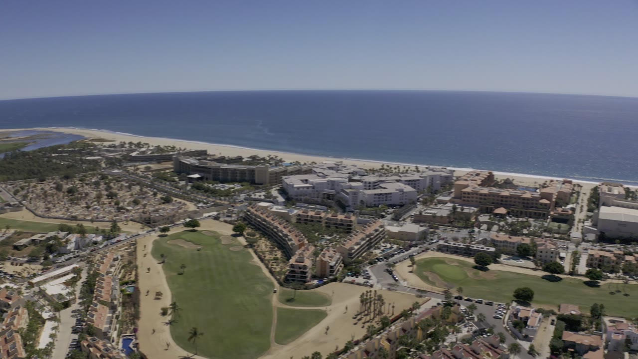 Wide aerial frame in Los Cabos, Mexico, showing the ocean horizon, sandy beach, apartment blocks, houses, roadways, and a green lawn area below, with no visible people.