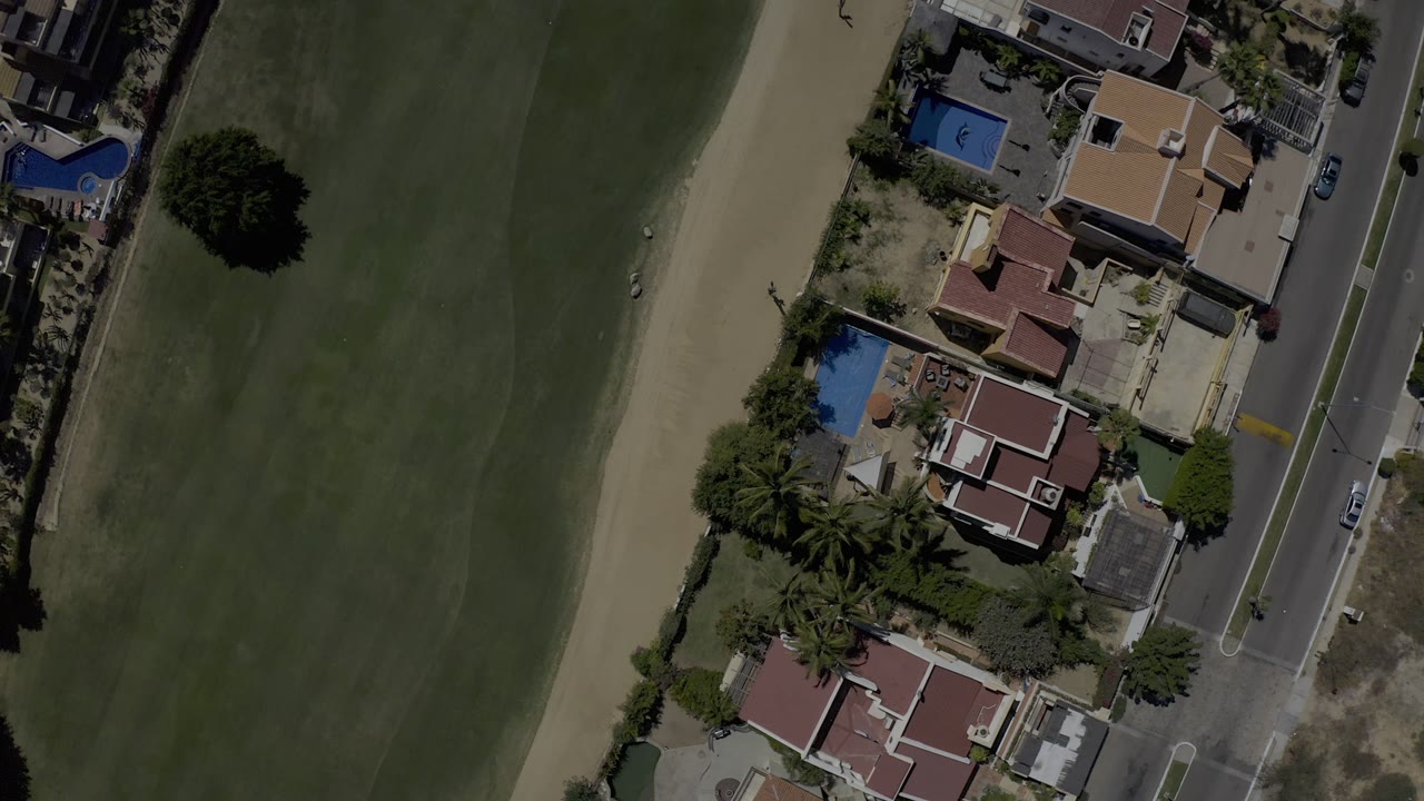 Near-vertical drone frame in Los Cabos, Mexico with residential roofs and backyard pools on one side of a road and a golf fairway on the other, with no visible people.