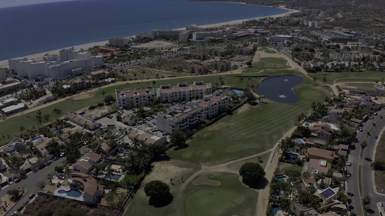 High aerial frame over Los Cabos, Mexico, showing residential buildings, golf course fairways, a small lake, beach, and blue sea with no clearly visible people.