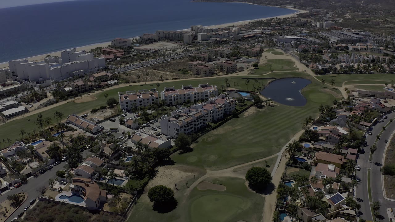 Los Cabos aerial view with apartment blocks and homes in the foreground, golf course greens and a lake in the center, and the beach and sea across the top of frame.