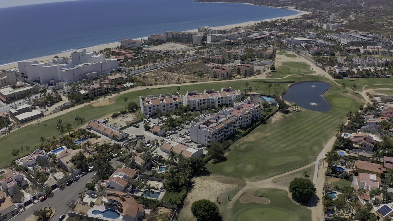 High aerial view in Los Cabos, Mexico, showing white coastal buildings, condominiums, golf greens, a pond, residential roofs, blue ocean, and no visible people.