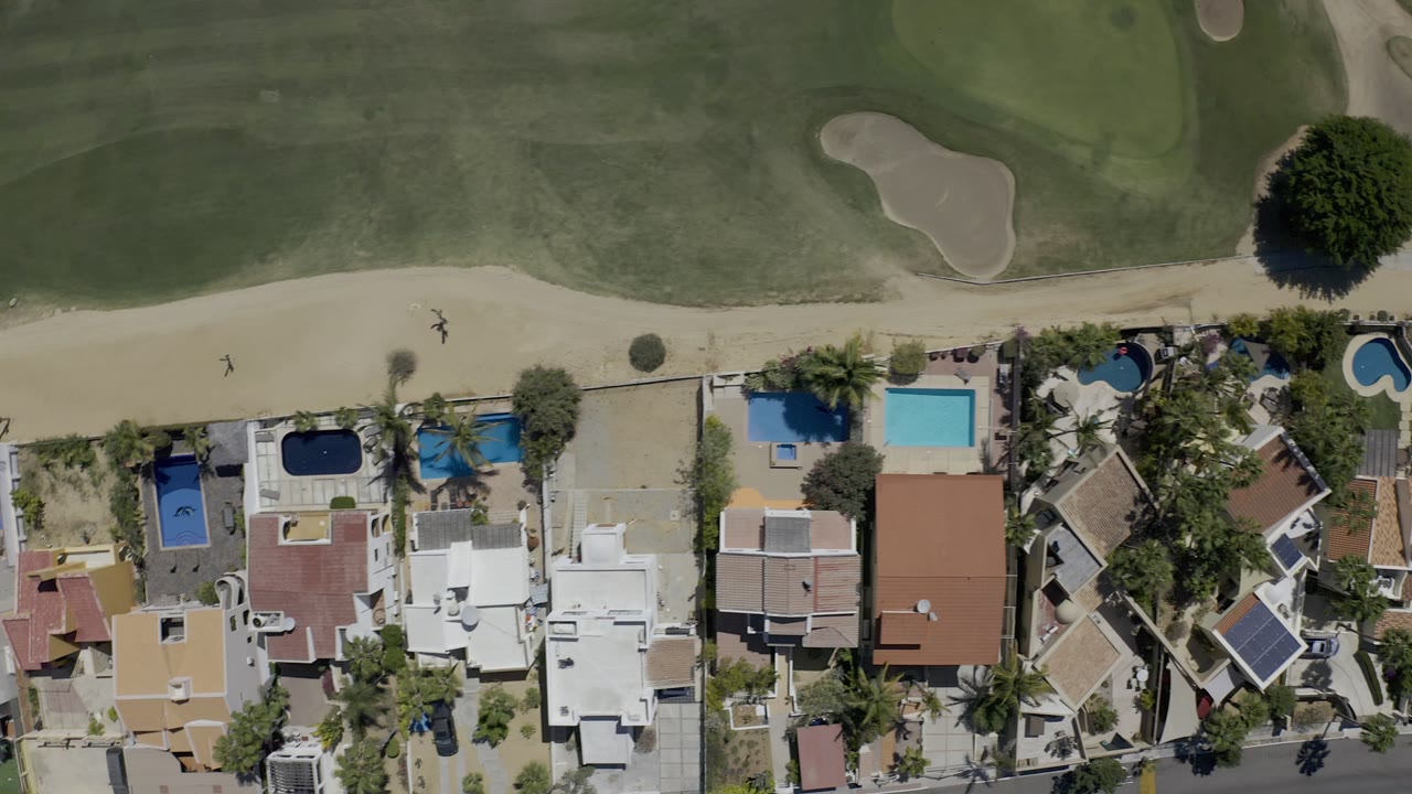 Aerial Los Cabos view with rooflines and blue pools along the lower frame and a curved sand path and fairway across the top, with no visible people.