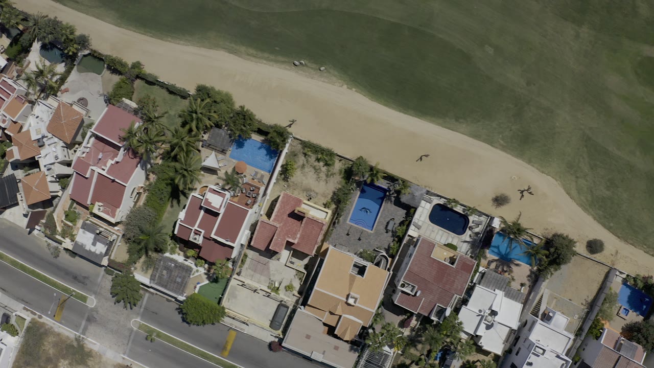Top-down aerial view of houses beside a road, pools, trees, sand, and green space.