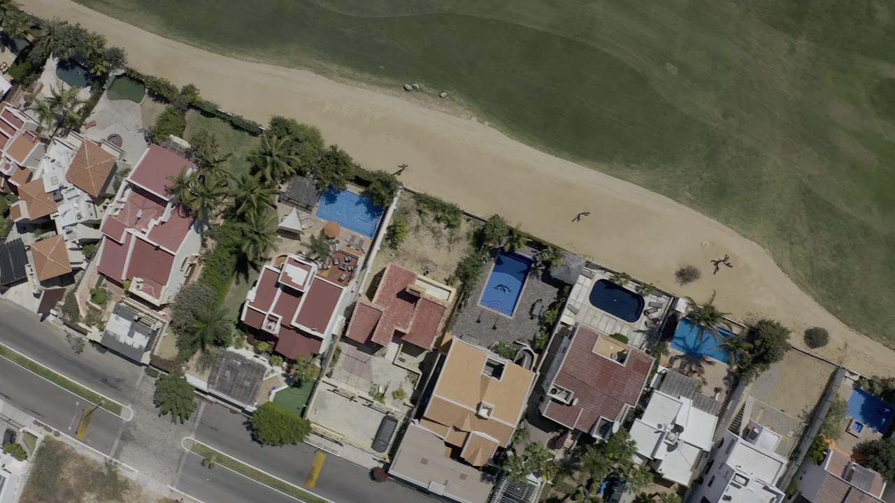Overhead Los Cabos frame with a row of private homes and courtyards along the lower edge, blue pools among palm trees, a road, and a broad green area above a pale sand strip with no visible people.