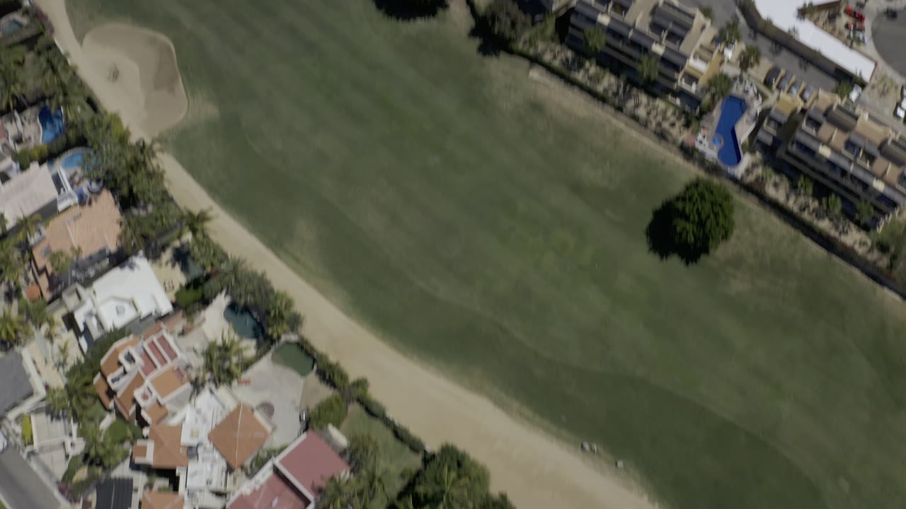 This Los Cabos aerial frame keeps the golf course prominent, with fairway grass, a sand bunker, clustered homes, and service roads visible below with no visible people.
