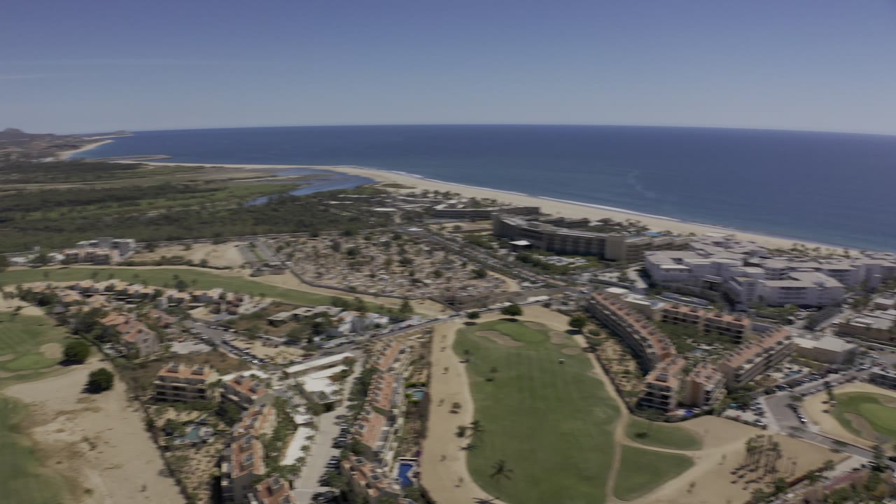 The next Los Cabos aerial view keeps the beach and blue ocean high in frame while resort blocks, streets, villas, and open green landscaping fill the lower view, with no visible people.