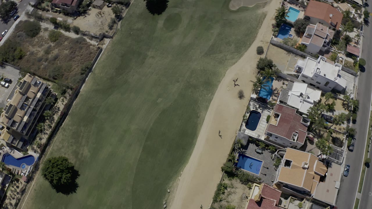 Los Cabos aerial frame looking down on a broad fairway bordered by a sand bunker, villas, pools, dry vegetation, and a straight road with no visible people.