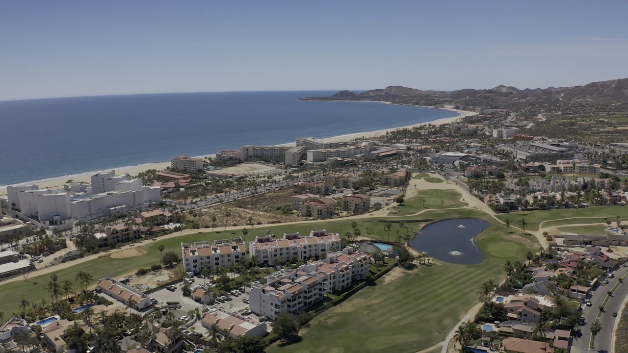 Wide aerial frame over Los Cabos, Mexico, with a golf course and pond in the foreground, white hotel buildings and rooftops near the beach, blue sea beyond, and no visible people.