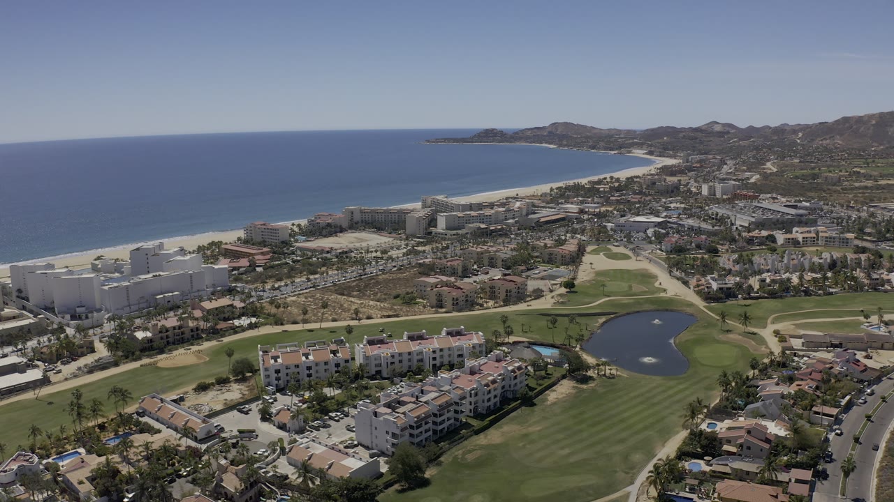 Aerial Los Cabos view showing fairways and a dark pond below, blocks of coastal buildings, a long pale beach curving beside blue water, distant hills, and no visible people.