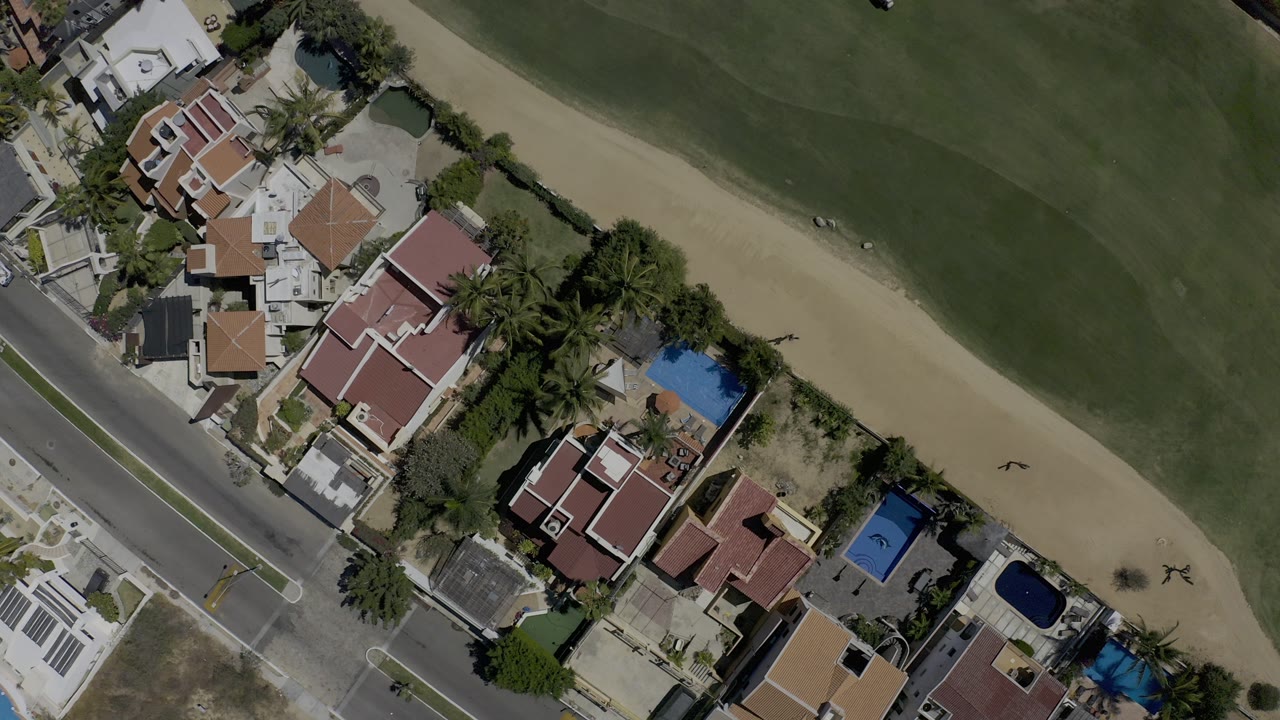 Near-vertical aerial view in Los Cabos, Mexico, showing red-roof homes, palm trees, a road, a green fairway, and sandy course areas with no visible people.
