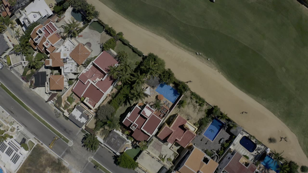 High aerial frame over Los Cabos, Mexico, with homes, a straight road, palms, green fairway, sandy cart paths or bunkers, and no visible people.