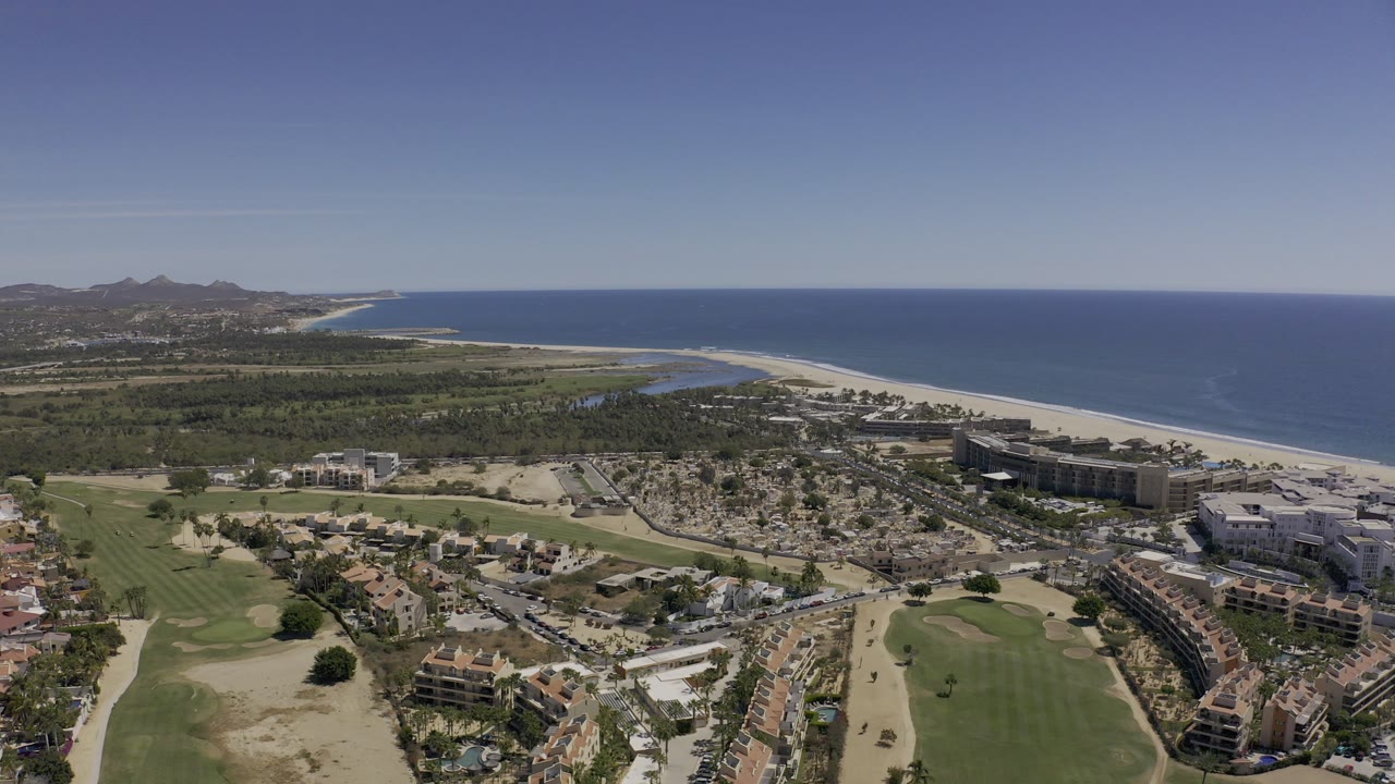 Wide aerial view over Los Cabos, Mexico with golf fairways in the foreground, shoreline buildings, a pale sand beach, blue Pacific water, distant hills, and no visible people.