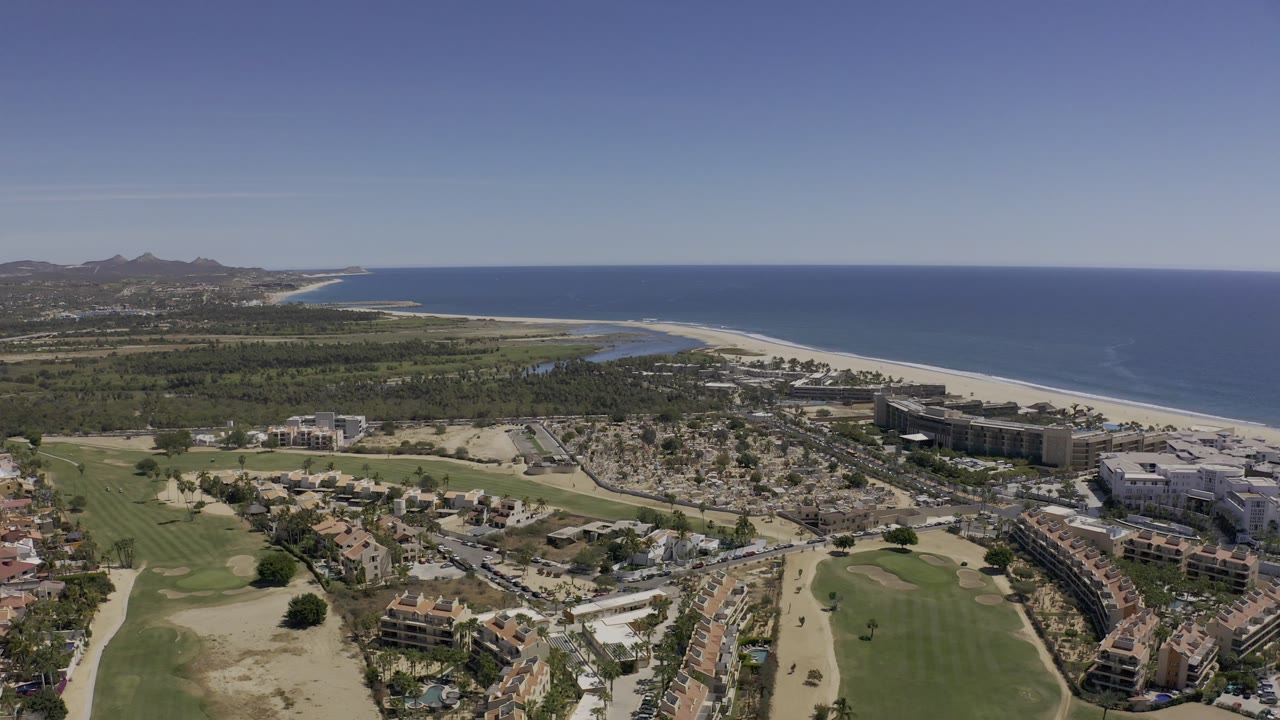Los Cabos aerial frame showing green golf terrain and desert ground below, low-rise shoreline buildings in the middle distance, a long sand beach, blue ocean, and no visible people.