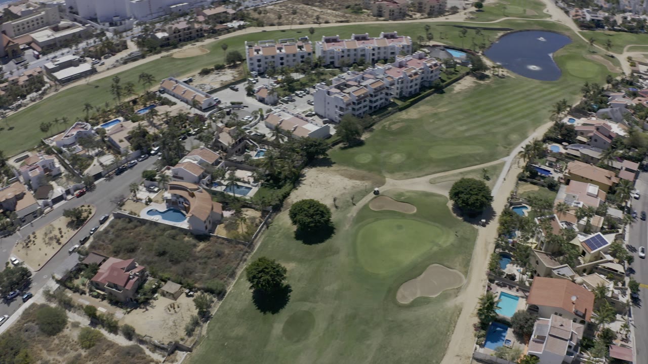 Aerial view in Los Cabos, Mexico, looking over residential homes, a golf green, curved streets, and a small pond near the coastline, with no visible people.
