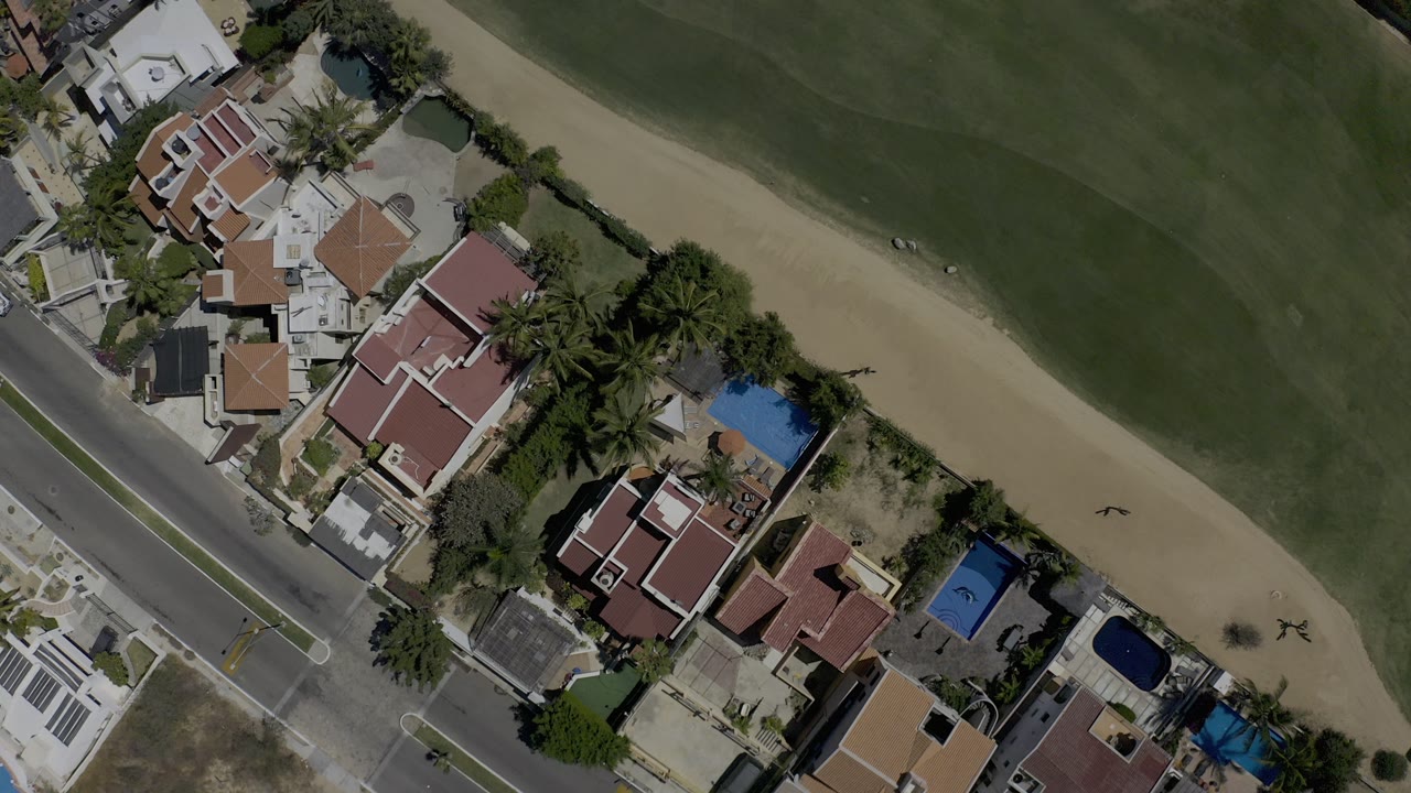 Overhead drone view in Los Cabos, Mexico, showing red-roofed homes, blue pools, a paved road, palm trees, and the edge of a golf fairway with no visible people.
