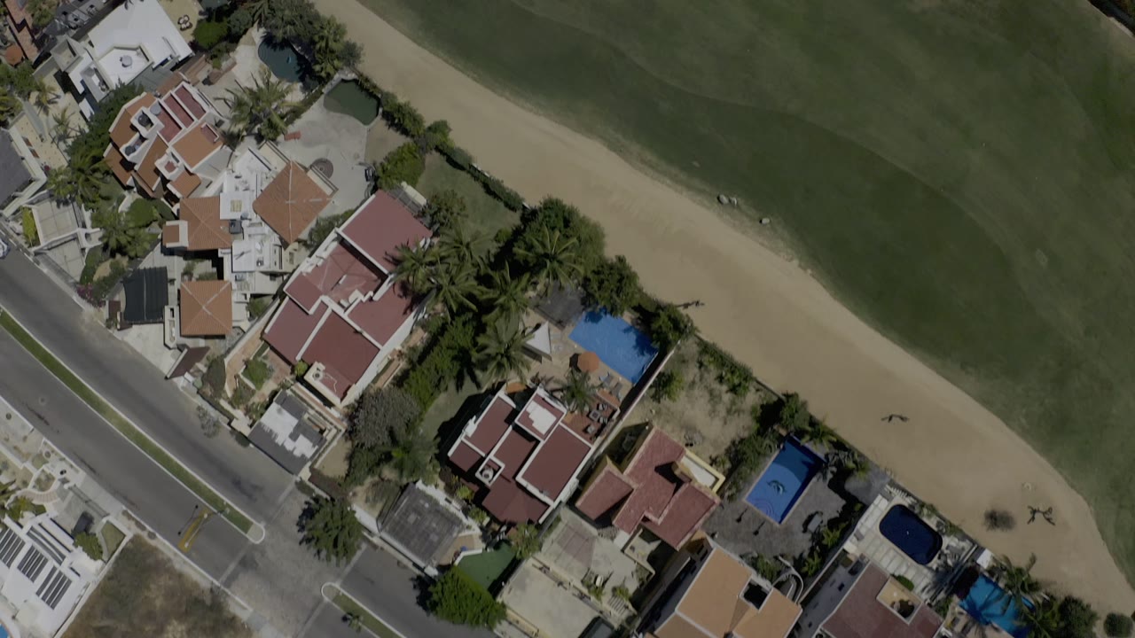 A slightly wider overhead view of Los Cabos homes, swimming pools, palm landscaping, a road, and a curved strip of golf-course green with no visible people.