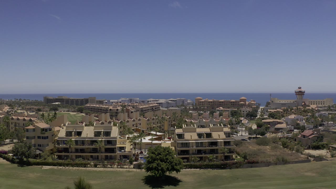 Wide aerial frame over Los Cabos, Mexico, with grass in the foreground, beige buildings and open lots below, blue water on the horizon, and no visible people.