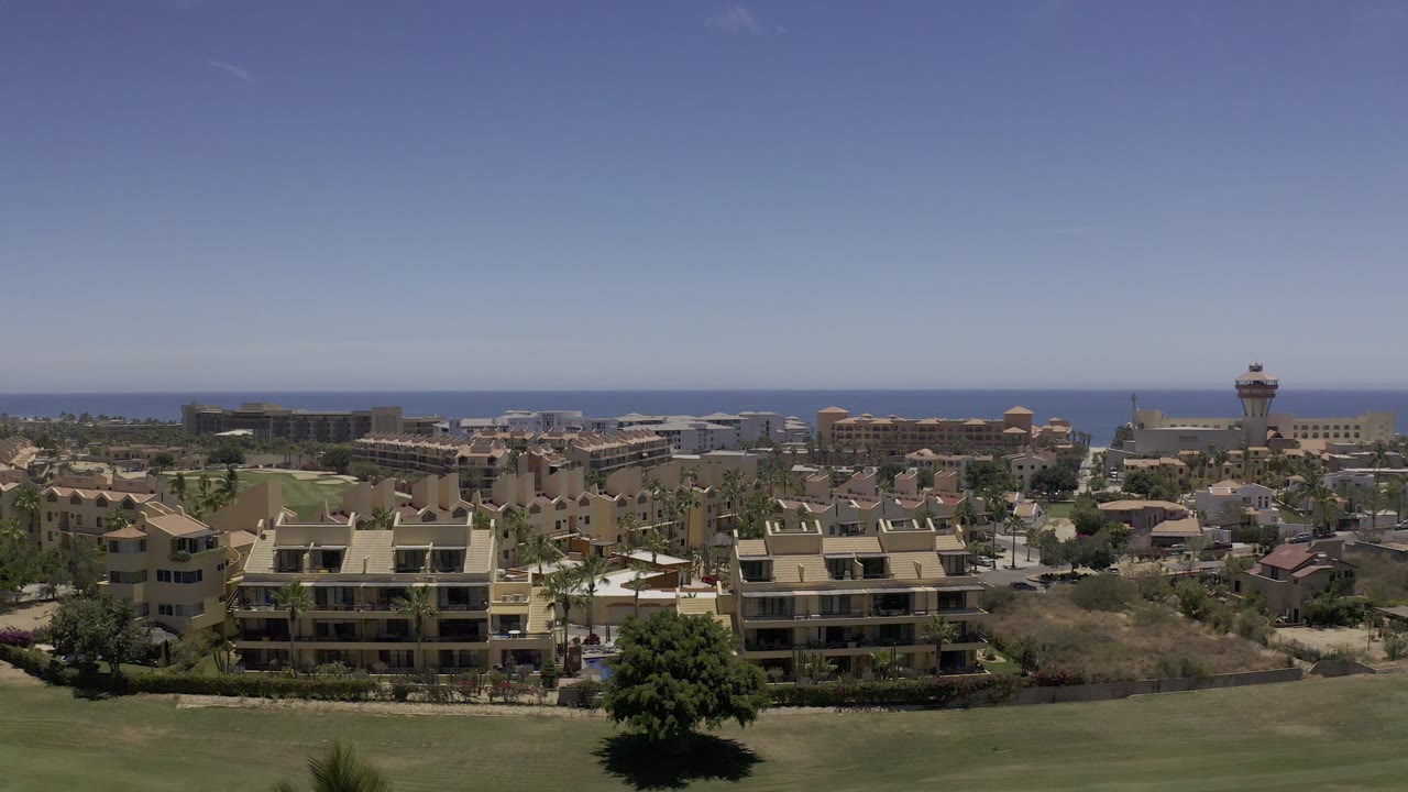 Los Cabos aerial view with a large beige residential-style building near the foreground, low-rise development across the midground, ocean in the distance, and no visible people.