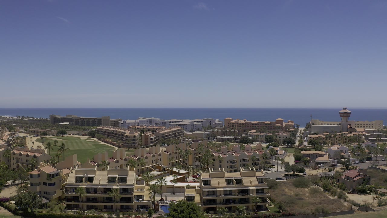 Opening overhead view of Los Cabos, Mexico, with tan residential buildings in the foreground, a green course or lawn at left, resort-style blocks beyond, the ocean on the horizon, and no visible people.