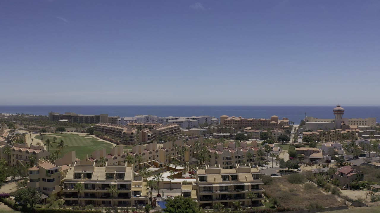 High aerial-style frame over Los Cabos, Mexico, showing apartment rooftops, a curving road, green open space, dense low-rise buildings, a distant ocean band, and no visible people.