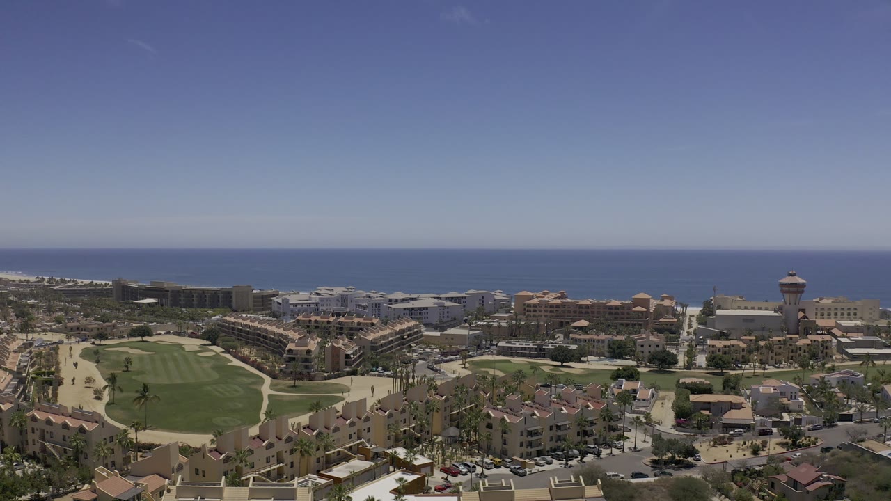 Aerial frame in Los Cabos, Mexico, showing the fairway on the left foreground, compact resort-area buildings in the middle distance, open sea beyond, and no visible people.
