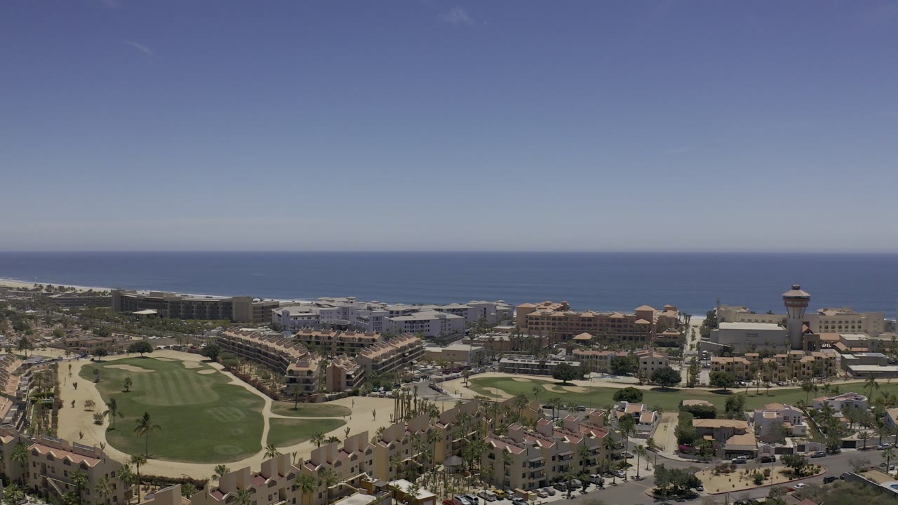 Aerial view of coastline, resort buildings, roads, and golf greens under a blue sky.