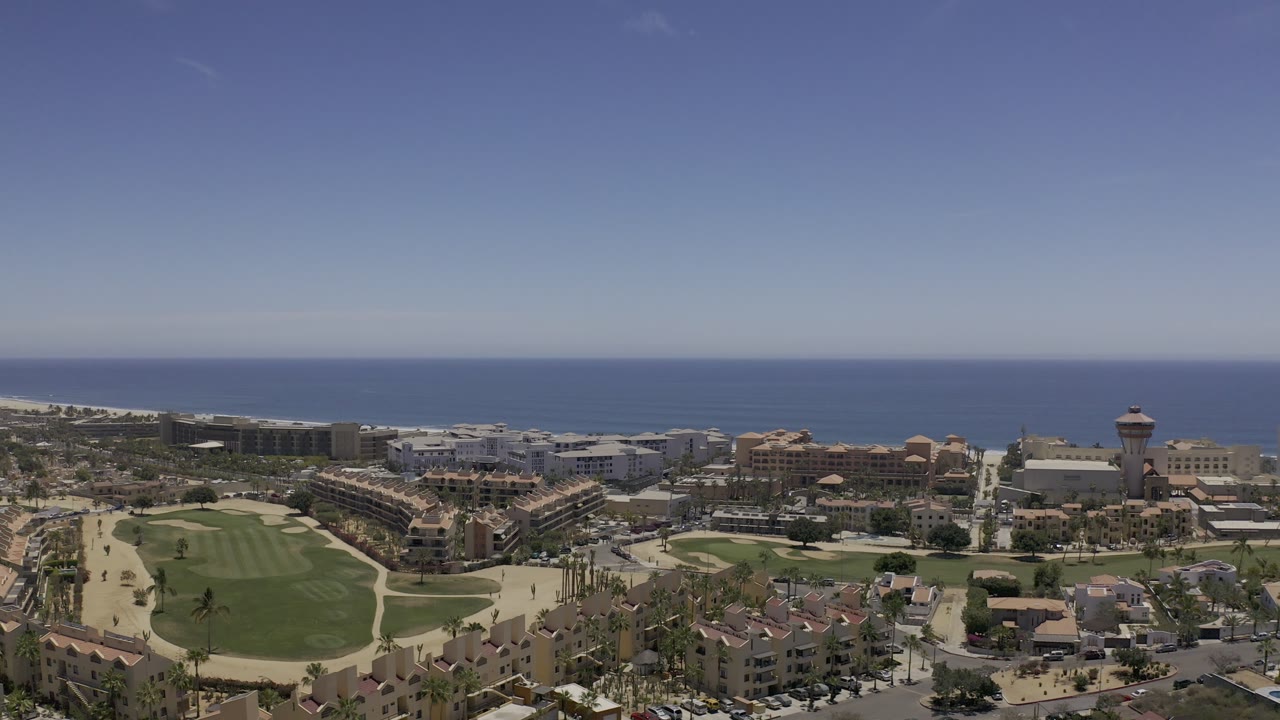 Aerial Los Cabos frame with fairways and sandy paths low in the image, dense buildings across the center, a tower-like structure to the right, and no visible people.
