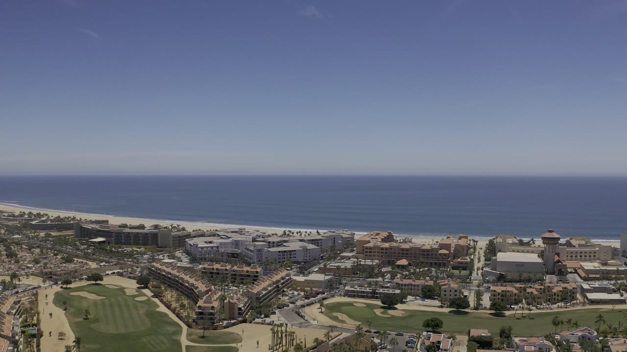Opening aerial frame over Los Cabos, Mexico, with golf greens in the lower frame, resort-style buildings near the shore, blue ocean on the horizon, and no visible people.