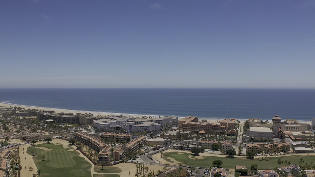 Los Cabos aerial view one second into the clip, showing green fairways at left, clustered coastal buildings in the center, sandy beach behind them, ocean beyond, and no visible people.