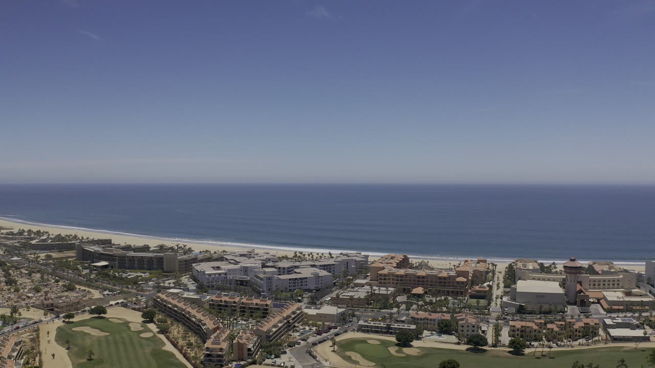 Wide aerial frame over Los Cabos, Mexico, showing city buildings below, a pale beach, blue ocean, and a large clear sky with no visible people.