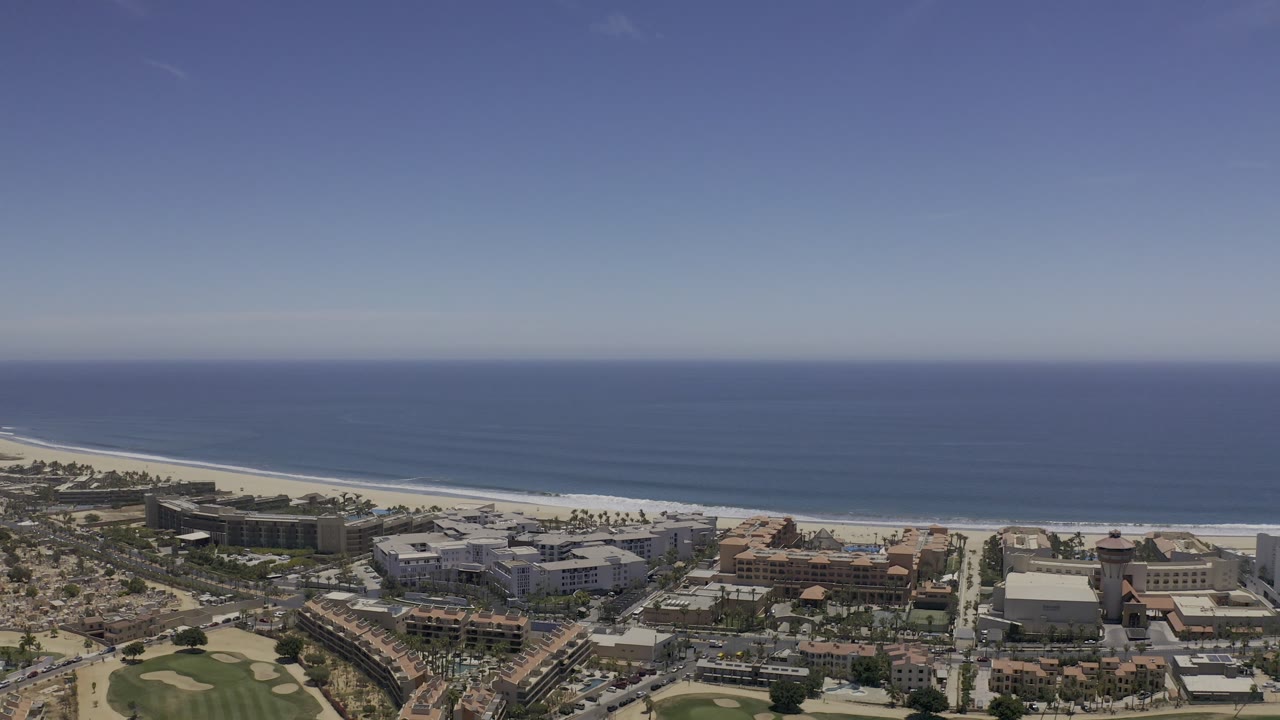 Early aerial view in Los Cabos, Mexico, showing the ocean horizon, white beach line, dense buildings, palm rows, and no visible people.
