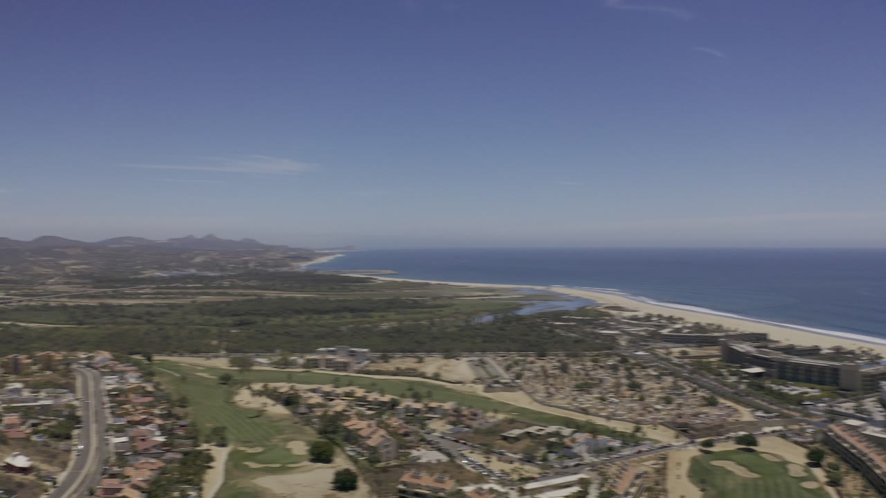 Aerial view of coastline, city roads, green areas, and distant hills under blue sky.