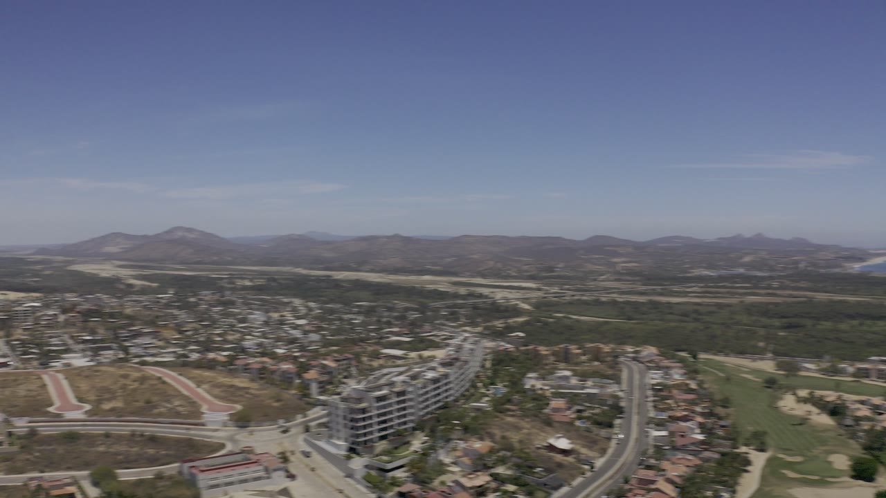 Los Cabos aerial looking inland across dense housing, paved roads, a track-shaped dirt area, modern buildings, dry hills, distant mountains, and no visible people.