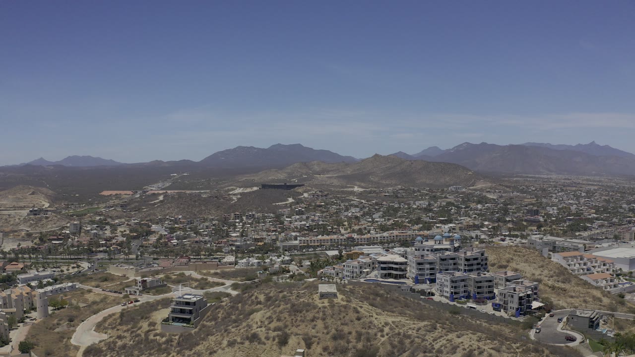 Opening aerial frame of Los Cabos showing scrub-covered hills in the foreground, modern low-rise buildings below, distant mountains, and no visible people.