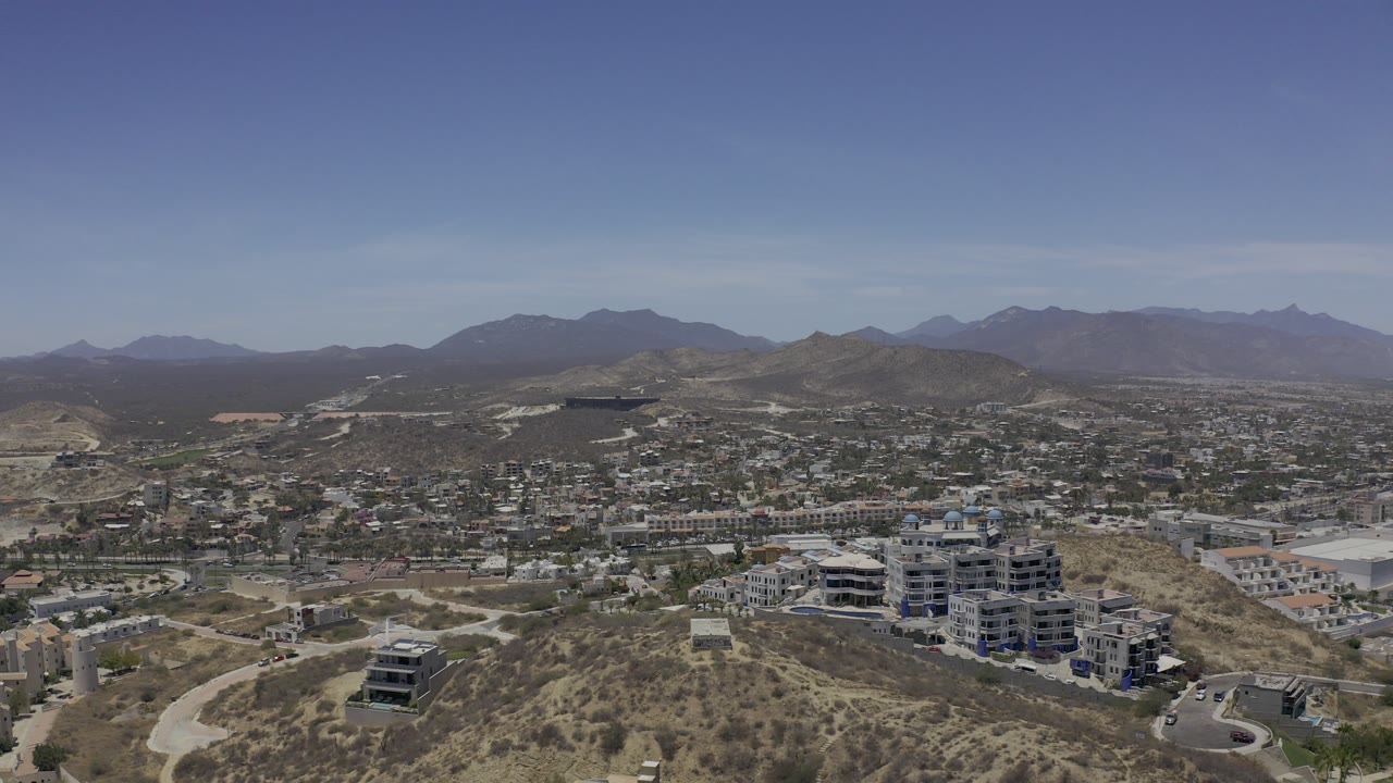 Aerial view over dry hills, roads, buildings, and distant mountains.