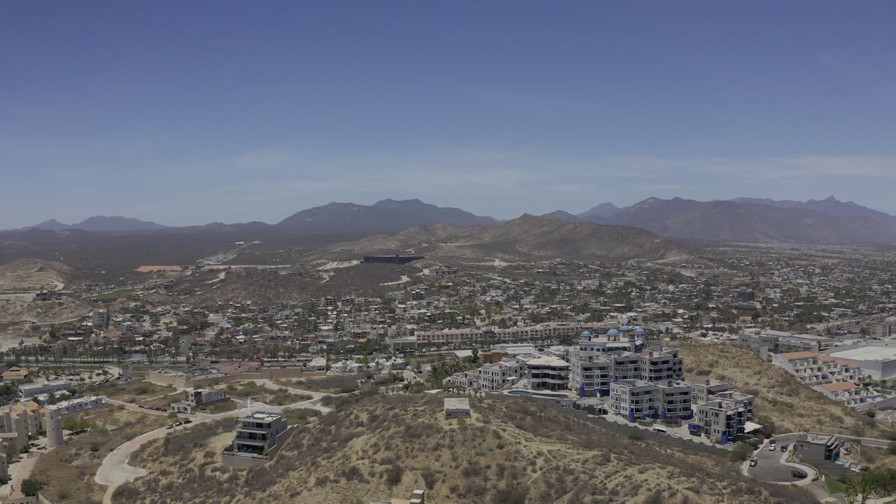 Wide aerial view in Los Cabos showing hillside construction, curving roads, scattered buildings, a broad mountain horizon, and no visible people.