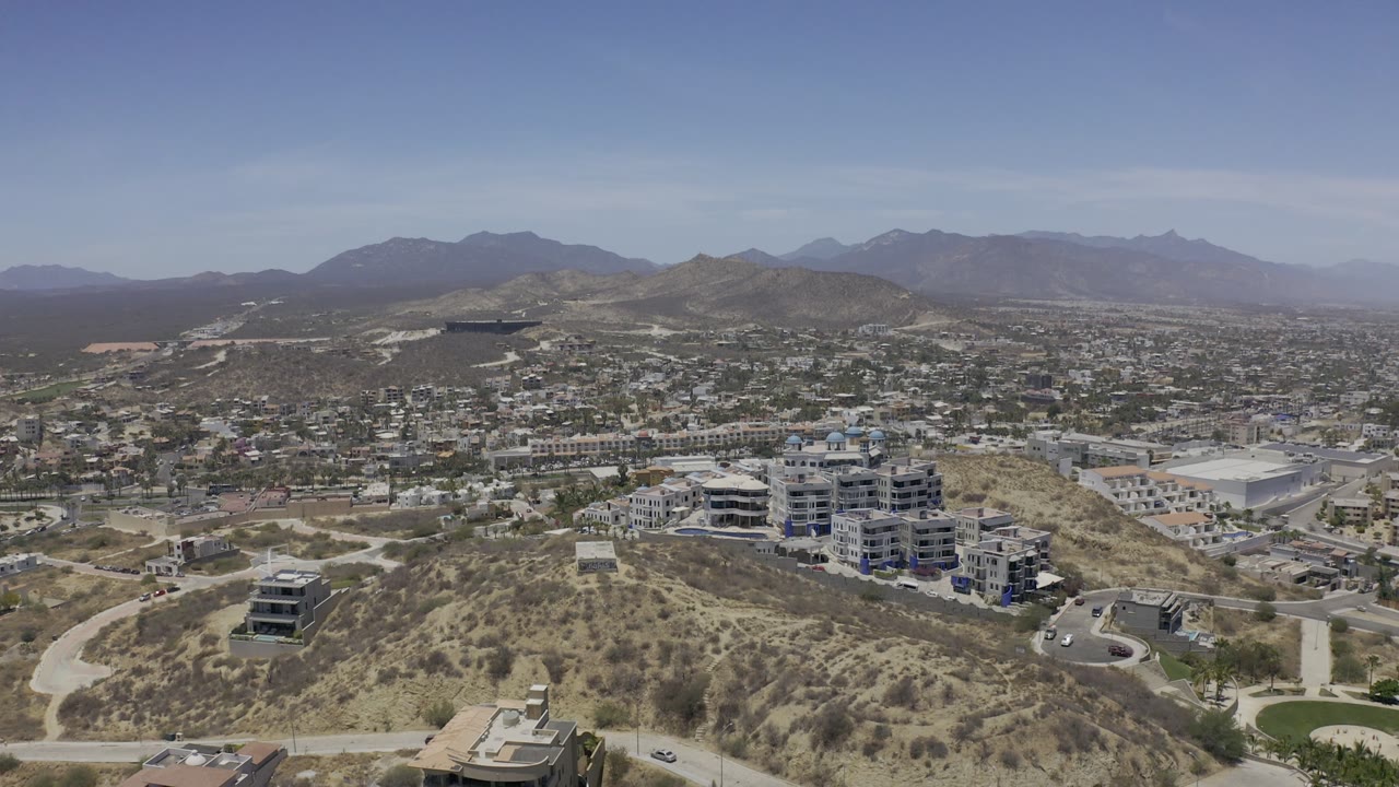 High aerial frame in Los Cabos, Mexico, with a dry hill in the foreground, modern buildings across the slope, roads below, hazy mountains, and no visible people.