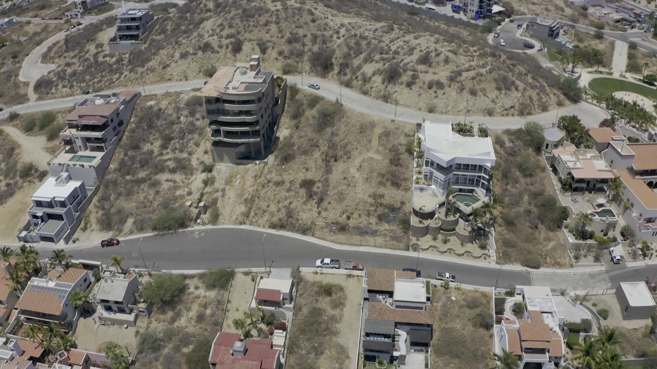High aerial view over Los Cabos, Mexico, showing modern hillside homes, dry scrub lots, a curved road, parked cars, and no visible people.